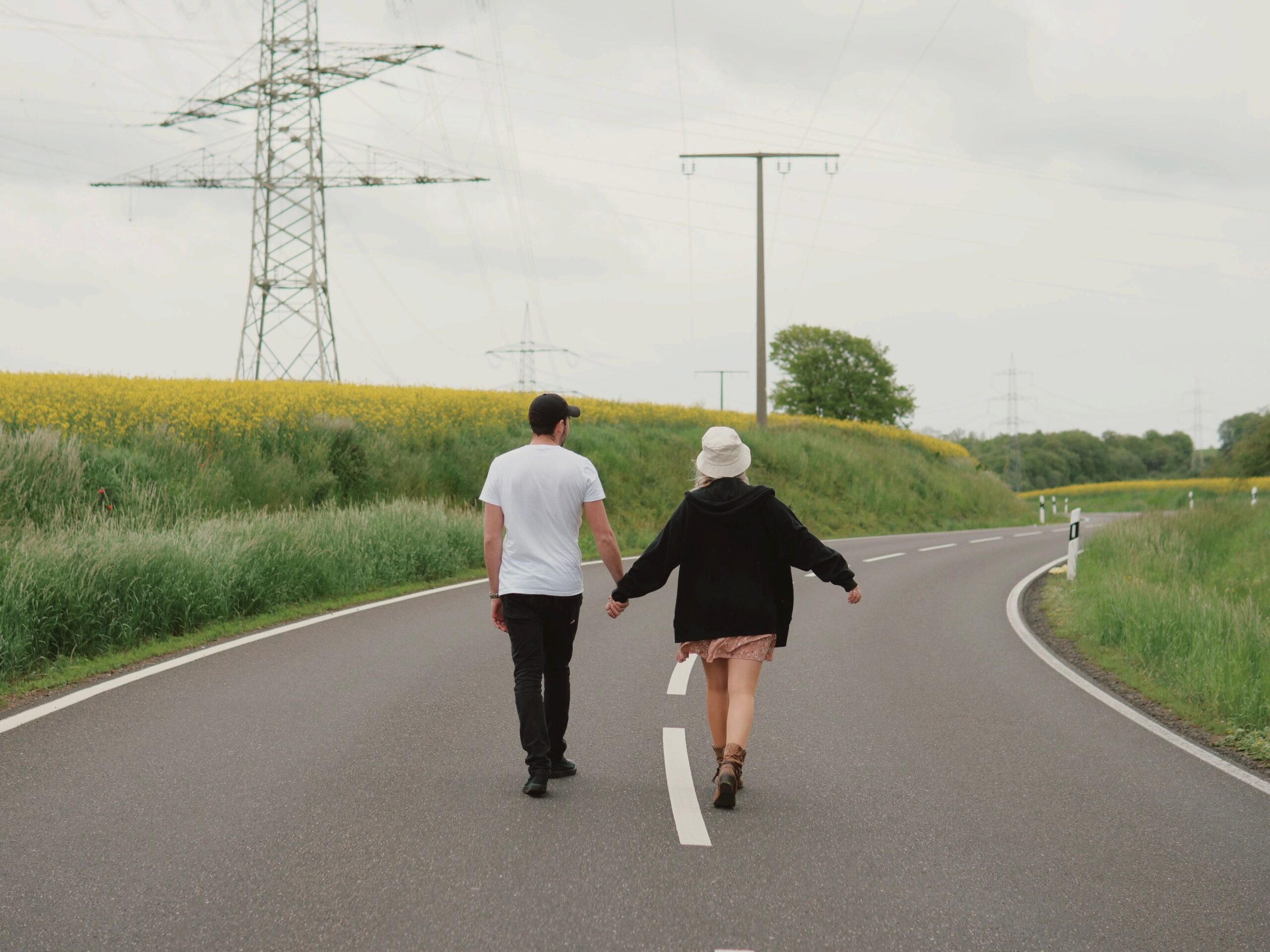A couple strolls hand-in-hand along a winding road surrounded by lush green fields.