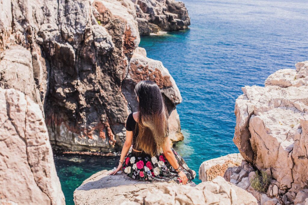 A woman sits on a rocky cliff, gazing at the clear blue Adriatic Sea in Croatia.